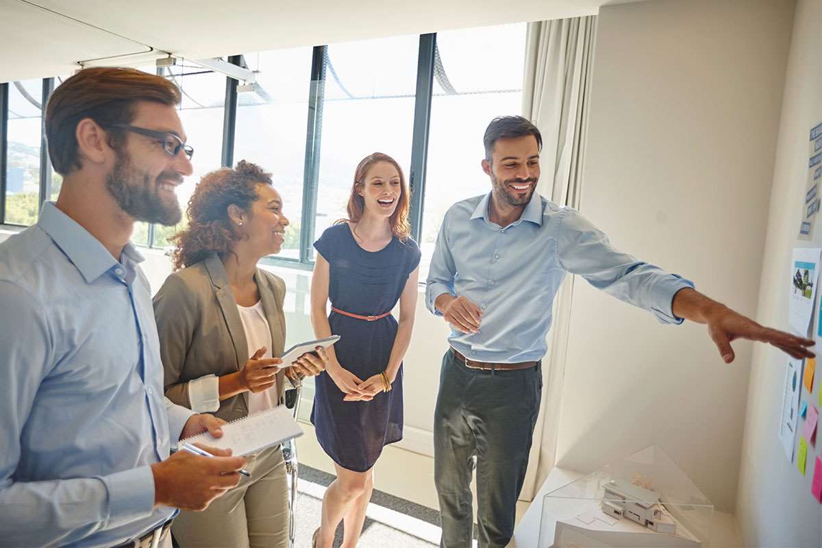 Two men and two women stand with one man gesturing toward some sticky notes on a wall in a brainstorming session.