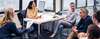Six team members sit around a office work station, with woman behind work station desk, collaborate on work