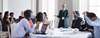 Standing woman and tables of people looking at woman and some people are raising their hand.
